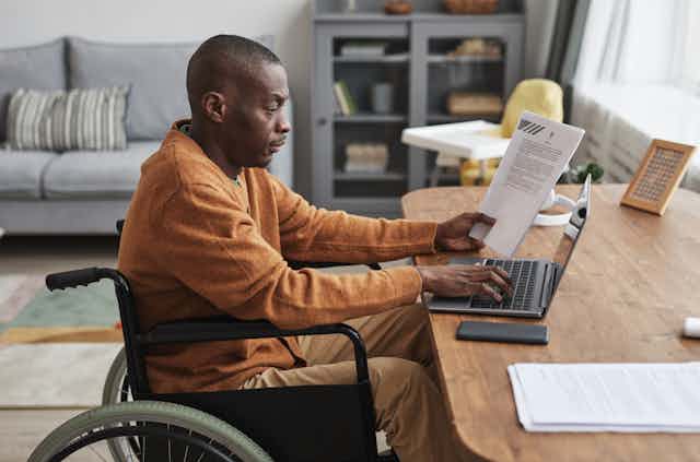 A Black man in a wheelchair works on a laptop at a table in a living room