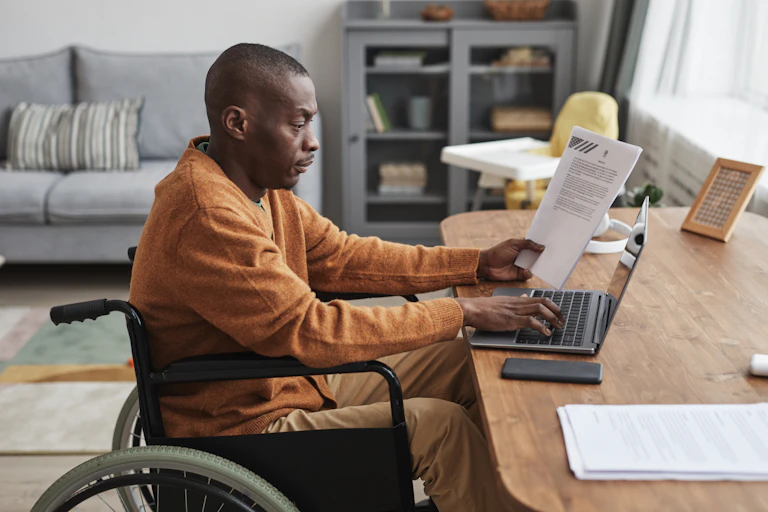 A Black man in a wheelchair works on a laptop at a table in a living room