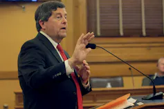 A dark-haired man speaks into a microphone.