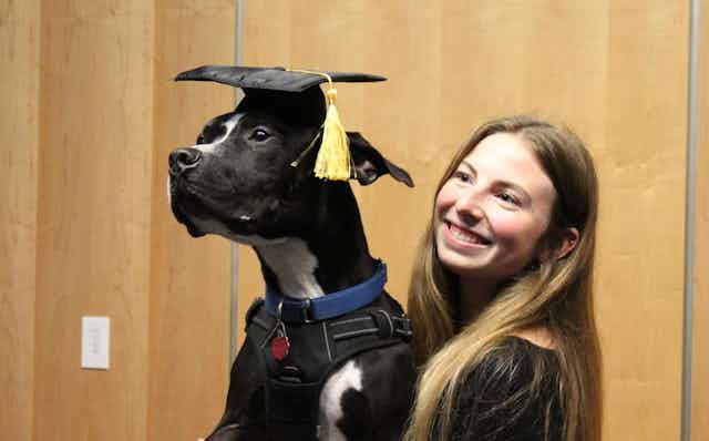 A young woman holding a dog, who is wearing a graduation cap with a yellow tassel hanging over its ear.