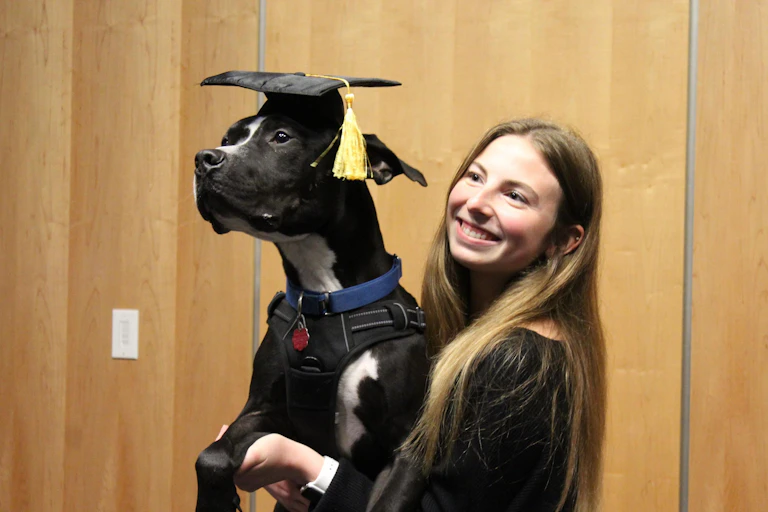 A young woman holding a dog, who is wearing a graduation cap with a yellow tassel hanging over its ear.