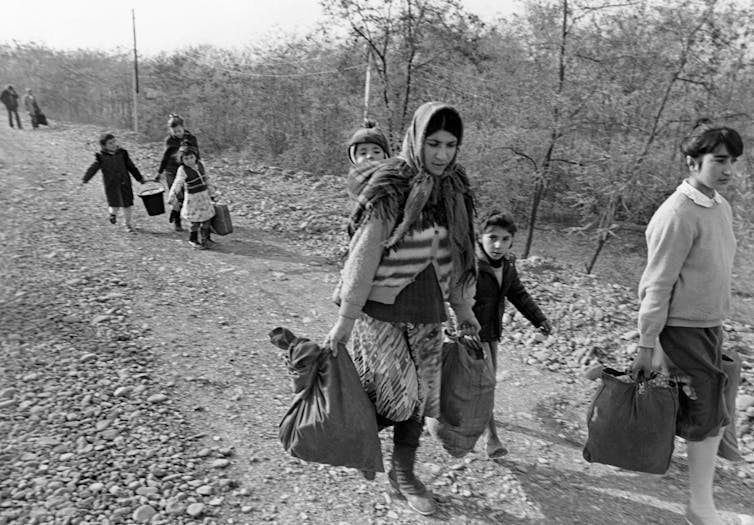 Women and children carrying bags walk down a dirt track.