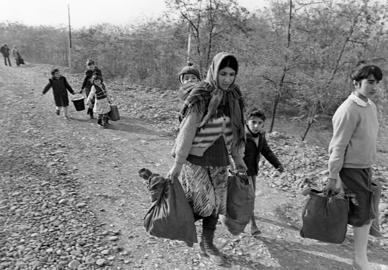 Women and children carrying bags walk down a dirt track.