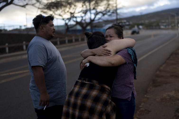 Dos supervivientes de los incendios de Lahaina se abrazan junto a una carretera mientras otro superviviente permanece a su lado.
