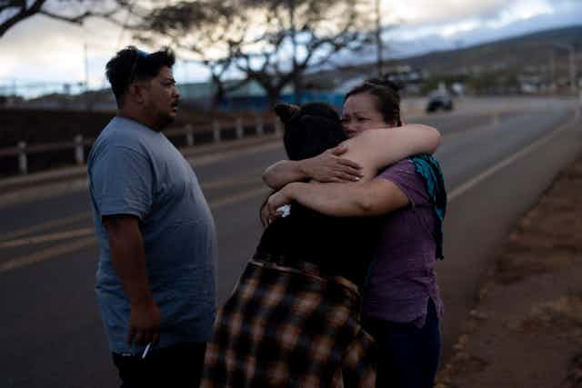 Two survivors of the Lahaina fires hug each other beside a road while another survivor stands beside them
