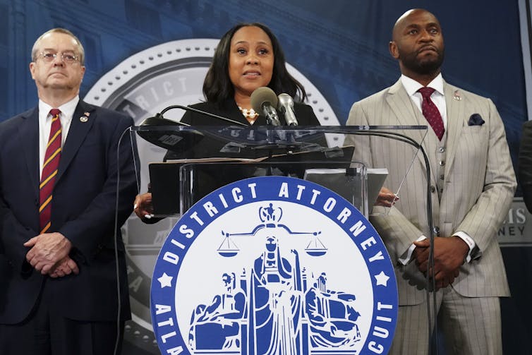 A woman in a dark jacket standing at a lectern that bears a seal that says 'District Attorney' across the top.