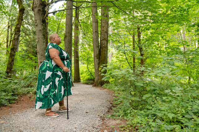 Side shot of a Black non-binary person standing in a rocky forest grove with their cane, looking upwards towards the surrounding greenery. They are in a flowing leaf print dress paired with gold sandals and have a shaved head, glasses, and tattoos above their elbow and ankle.