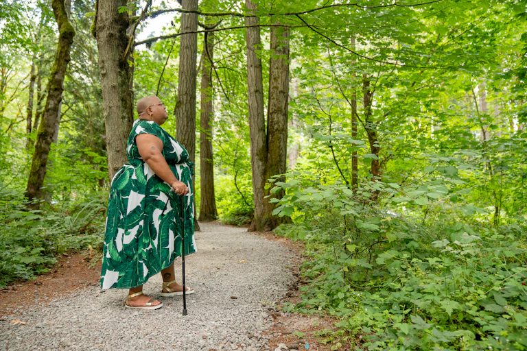 Side shot of a Black non-binary person standing in a rocky forest grove with their cane, looking upwards towards the surrounding greenery. They are in a flowing leaf print dress paired with gold sandals and have a shaved head, glasses, and tattoos above their elbow and ankle.