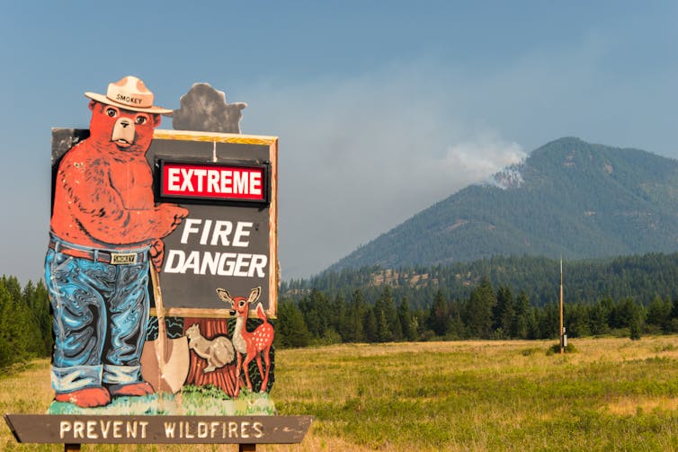 Pancarte avertissant sur le danger des feux de forets, avec derrière une montagne et de la fumée