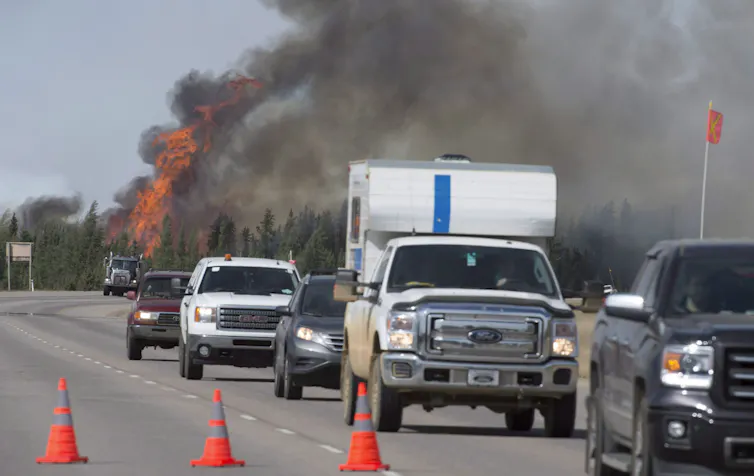 a row of cars on a road drives past fire and smoke in the background