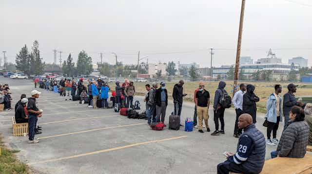 a row of people with packed bags wait in a carpark, the air is hazy with smoke
