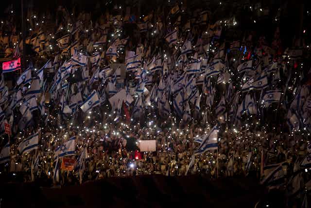 Thousands of people in the night in a crowd, many holding blue and white flags.