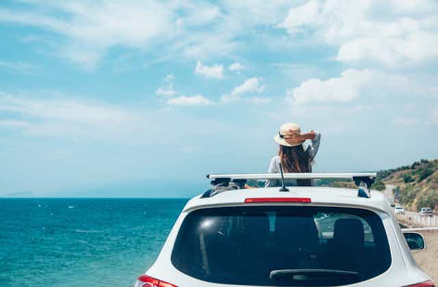A girl stands in a car, looking out the roof window at a beach.
