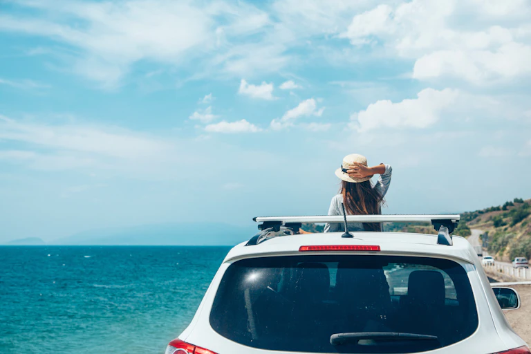 A girl stands in a car, looking out the roof window at a beach.