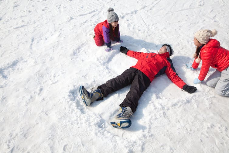 A man makes a snow angel in the snow, surrounded by a child and a woman.