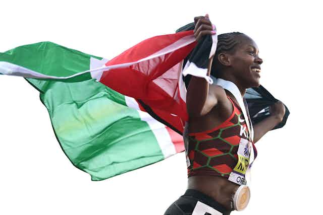A woman beams as she runs with a Kenyan flag held up in her arms behind her.