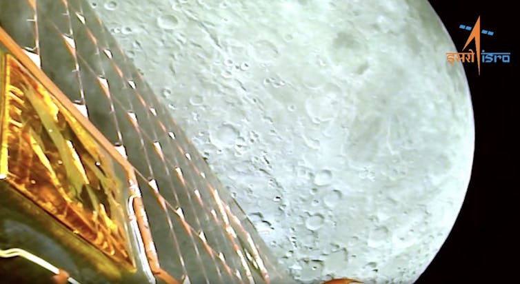 Surface of the moon seen past a solar array of orbiting spacecraft, with ISRO logo.