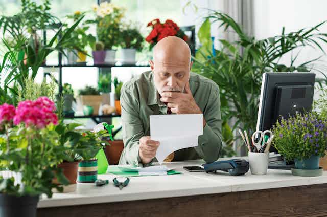 Man behind desk with computer, card reader, plants and floristry tools, looking at a piece of paper, worried expression.