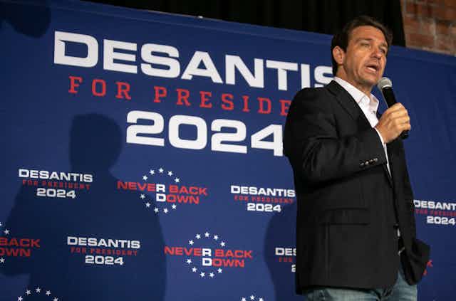 A dark-haired man in a dark jacket speaks into a microphone on a stage. DeSantis for President signs are behind him.