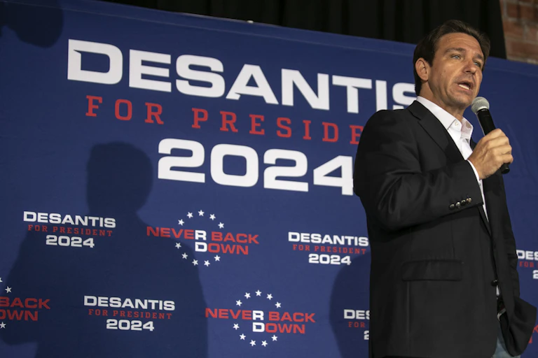 A dark-haired man in a dark jacket speaks into a microphone on a stage. DeSantis for President signs are behind him.