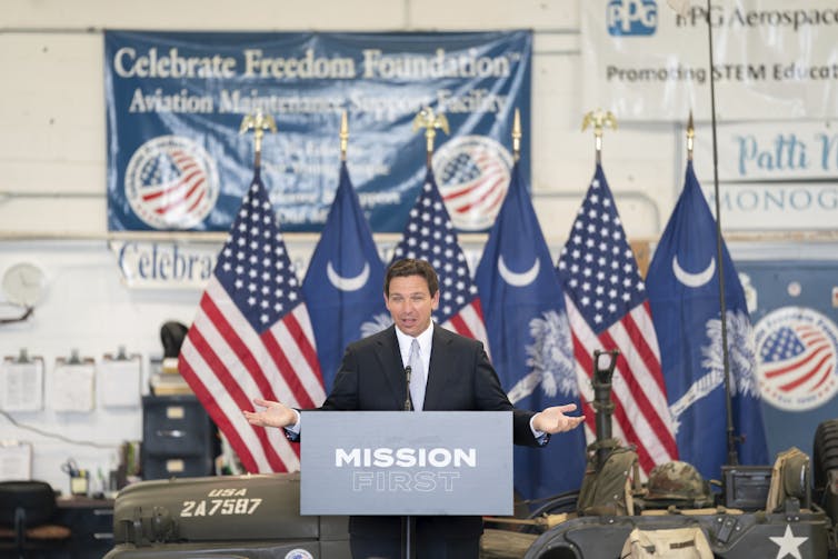 A dark-haired man speaks at a podium with American flags behind him.