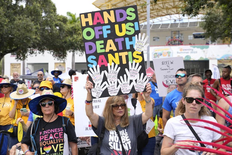 A woman in sunglasses holds a sign that reads Hand Off Us DeathSantis.