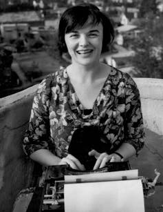 Black and white photograph of woman sitting on balcony smiling and using a typewriter.