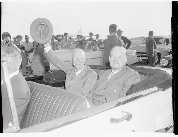 Dwight Eisenhower is one of two men shown in an open-top car in a black and white photo. He waves his hat in the air at a crowd of people.