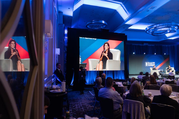 Nikki Haley is seen sitting on a stage and speaking, as seen from multiple television screens in a dark roo.