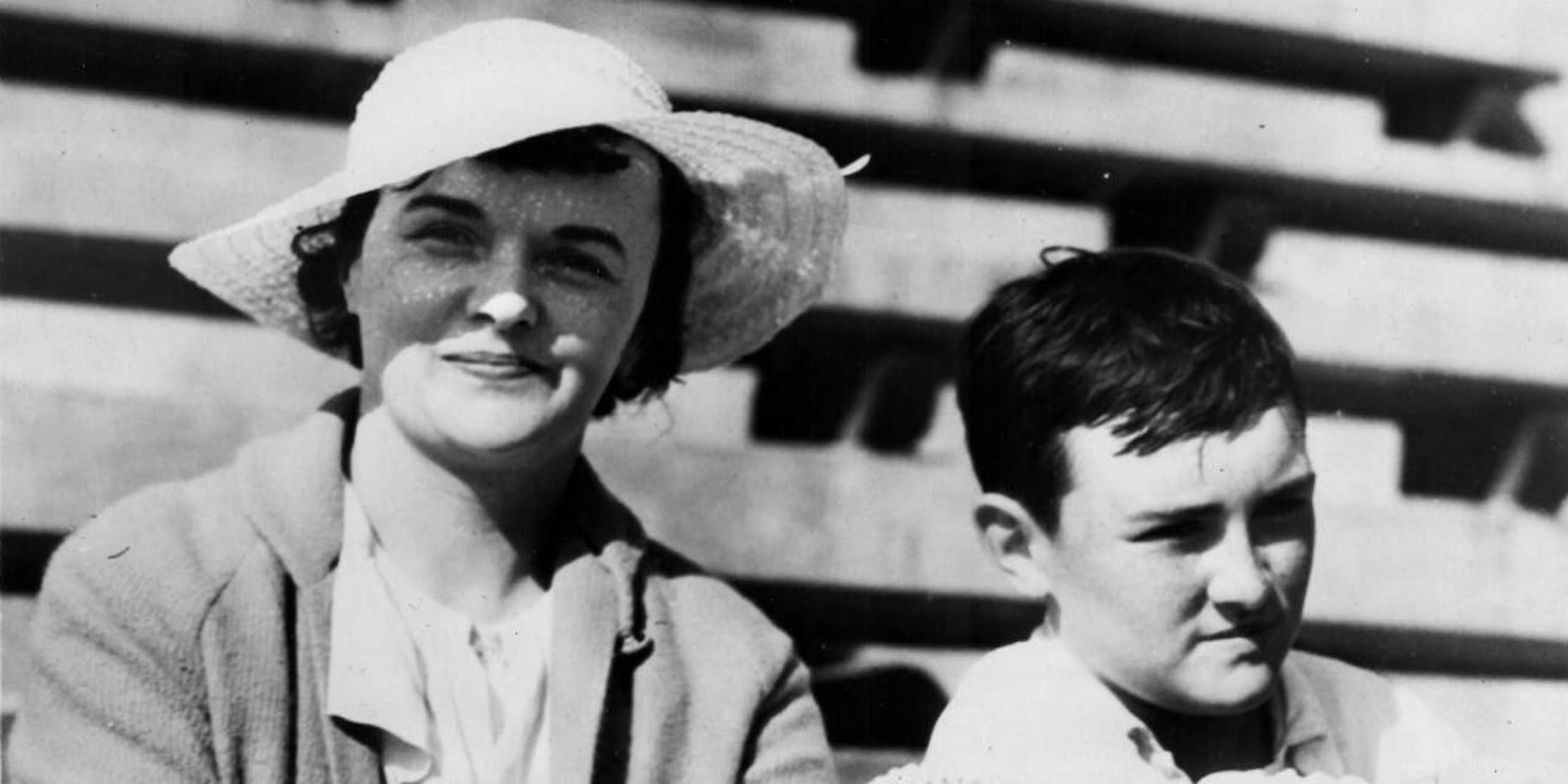 Black and white photo of woman wearing hat posing with boy in bleachers.