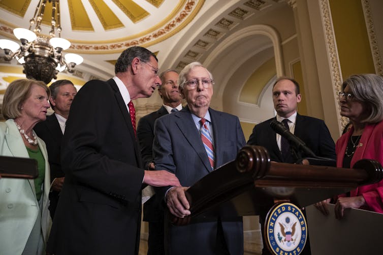 People in formal clothing gather around Mitch McConnell, who is at a lectern