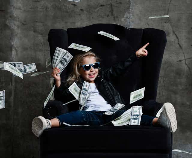 A girl sitting in an armchair holds a stack of banknotes in her hand while more banknotes fly around her.