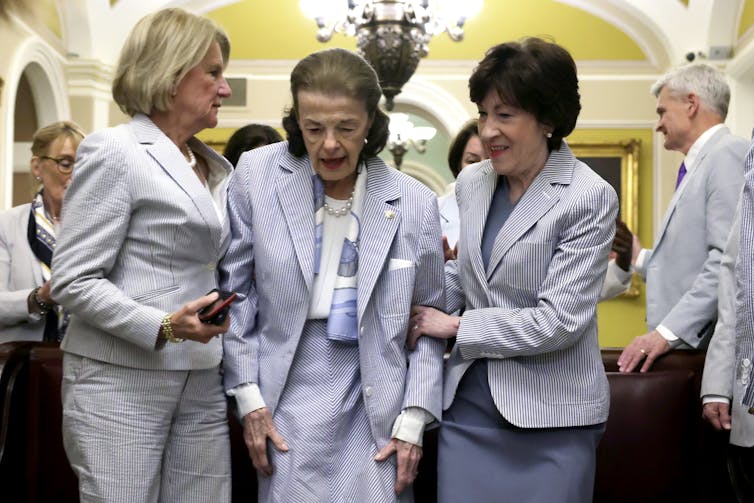 Diane Feinstein wears a light pantsuit, and is held up by two colleagues, both female, on either side.