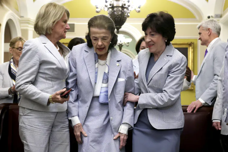 Diane Feinstein wears a light pantsuit, and is held up by two colleagues, both female, on either side.