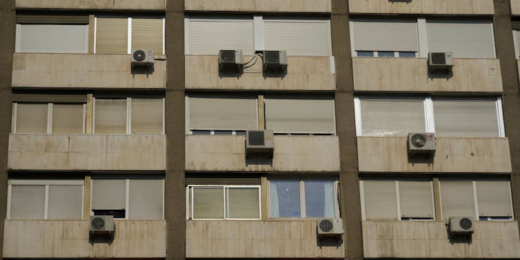 An apartment building wall with closed windows, an AC unit in each.