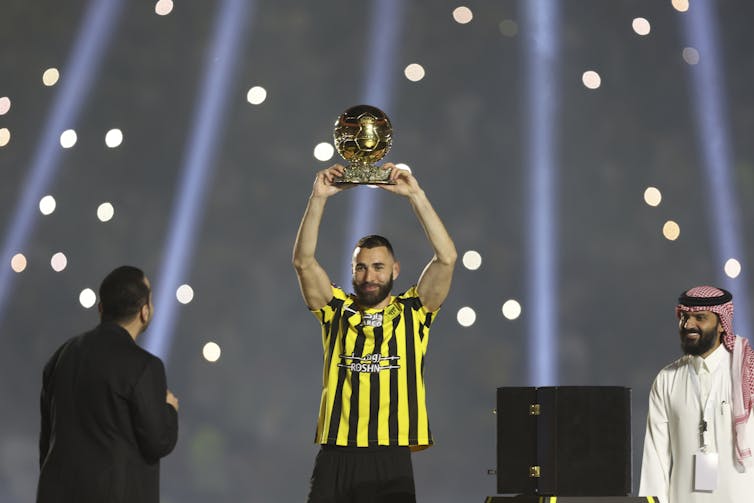 A man in a black and yellow football shirt raises a golden trophy above his head.