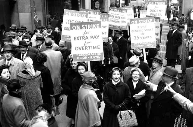 Men and women in dressy winter attire mill around carrying signs indicating they are on strike in a black and white photo from the 1950s.