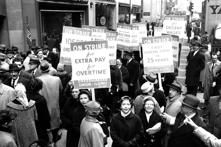 Men and women in dressy winter attire mill around carrying signs indicating they are on strike in a black and white photo from the 1950s.