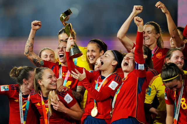 A group of women in red soccer jerseys cheer while wearing gold medals. One women in the middle holds a trophy up