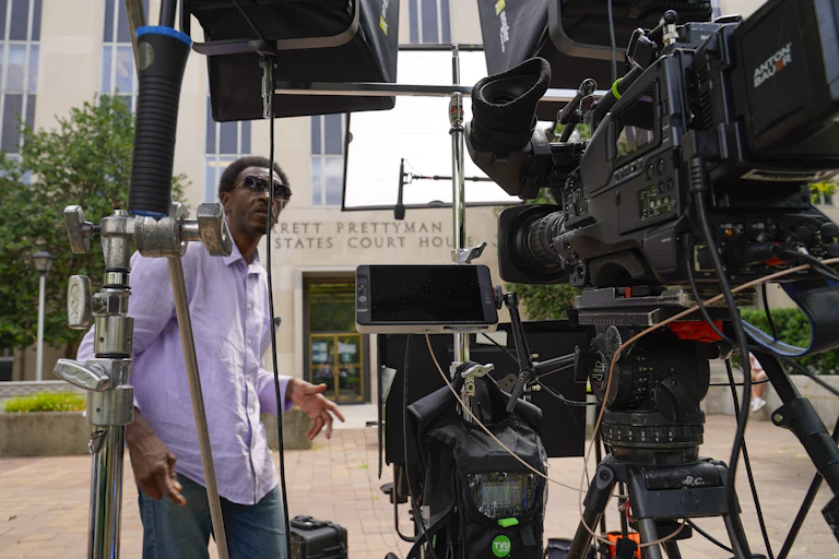 A man in a purple shirt standing among a lot of camera equipment outside a large building.