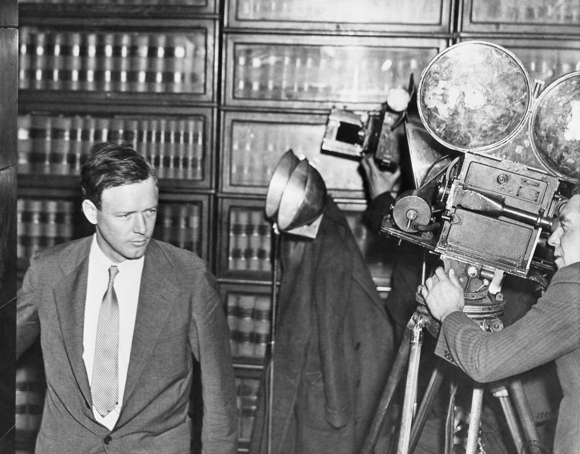 A man in a suit in front of bookshelves being filmed by old-fashioned cameras.