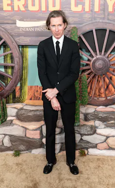 A man in a black suit standing in front of a backdrop of wagon wheels and cacti.