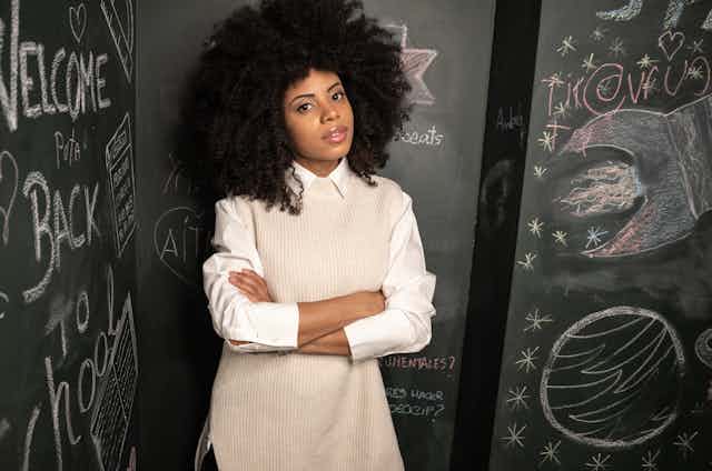 A confident Black woman with an afro stands with her arms folded near a chalkboard with the words