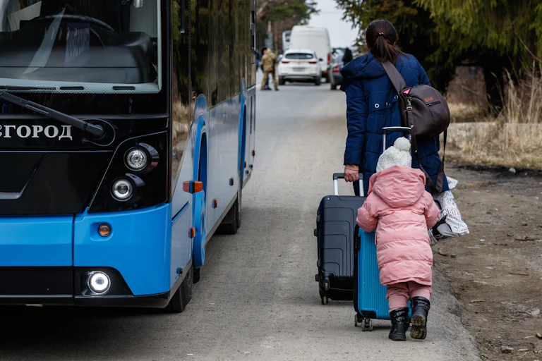 View from behind of a woman and a small child behind her, pushing large suitcases as they walk along the side of a road