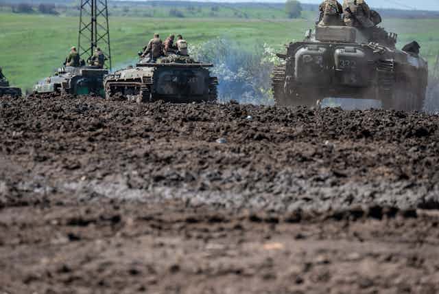 Three tanks with soldiers on top, riding through a muddy field.