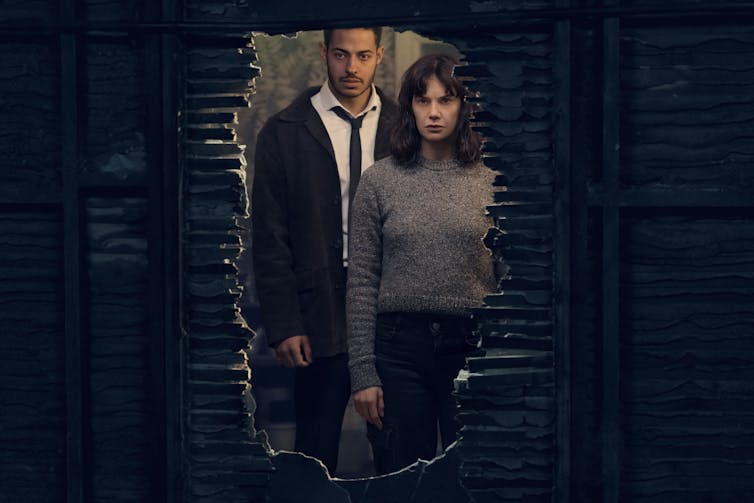 Lorna and the detective look through a hole in a wall.