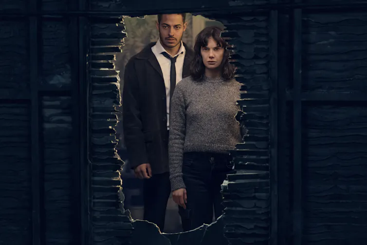 Lorna and the detective look through a hole in a wall.