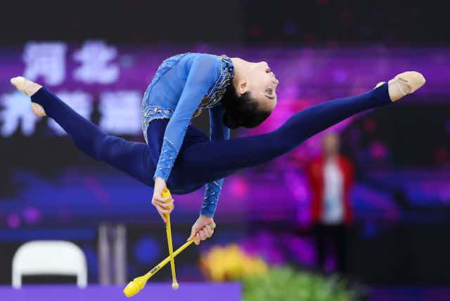 A rhythmic gymnast leaps in the air with a pair of yellow sticks. Her back arches so her head nearly touches the back of her thighs.