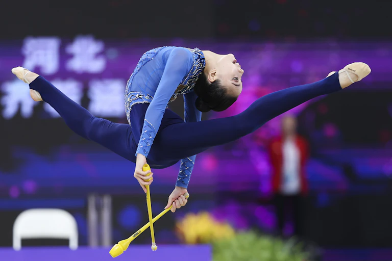 A rhythmic gymnast leaps in the air with a pair of yellow sticks. Her back arches so her head nearly touches the back of her thighs.