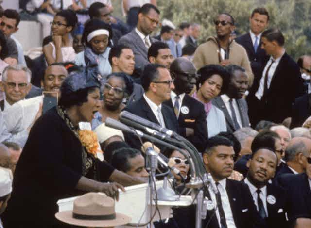 A Black woman is singing in front of several microphones as mostly Black people sit and listen. 